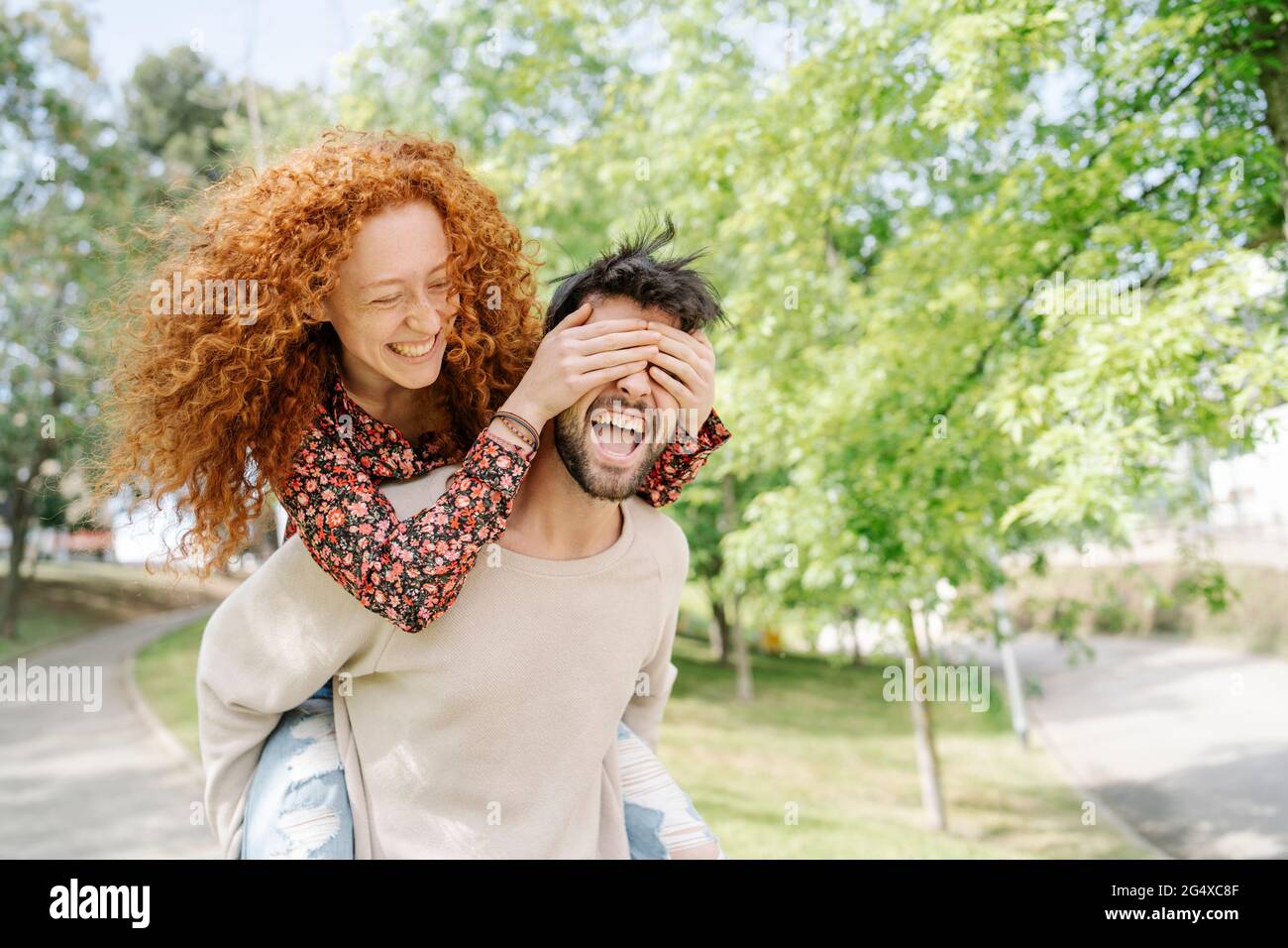 Cheerful boyfriend carrying girlfriend piggyback while covering eyes in park Stock Photo