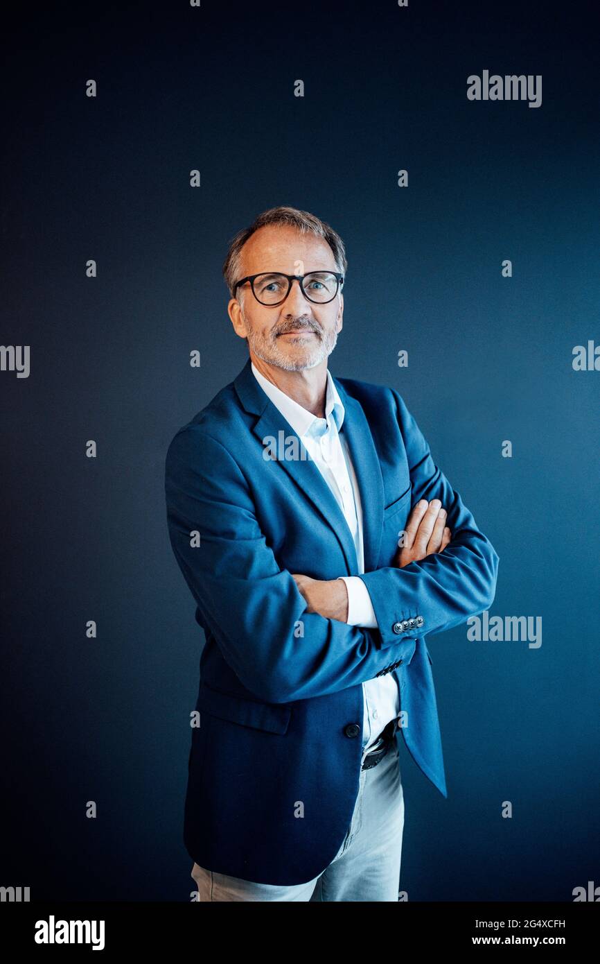 Confident businessman standing with arms crossed in front of blue wall ...