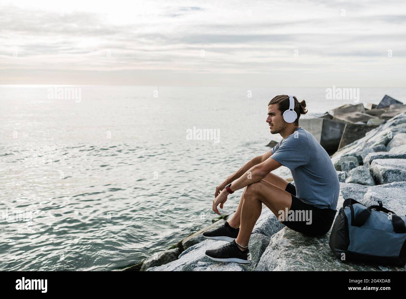 Sportsman with headphones looking at view while sitting on rock by sea Stock Photo