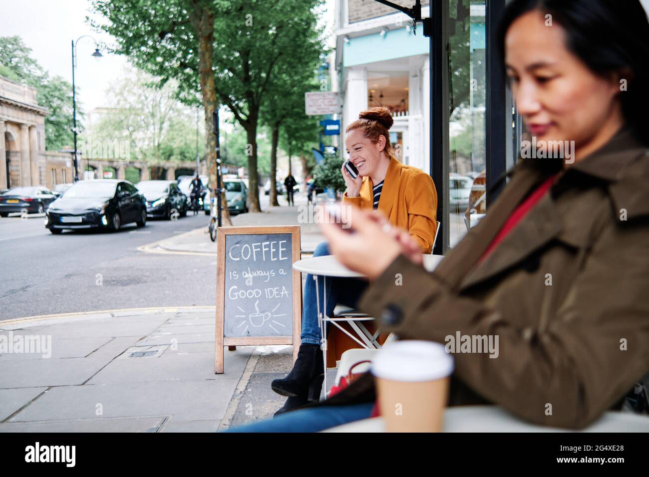 Smiling woman talking on smart phone with female friend sitting at sidewalk cafe Stock Photo