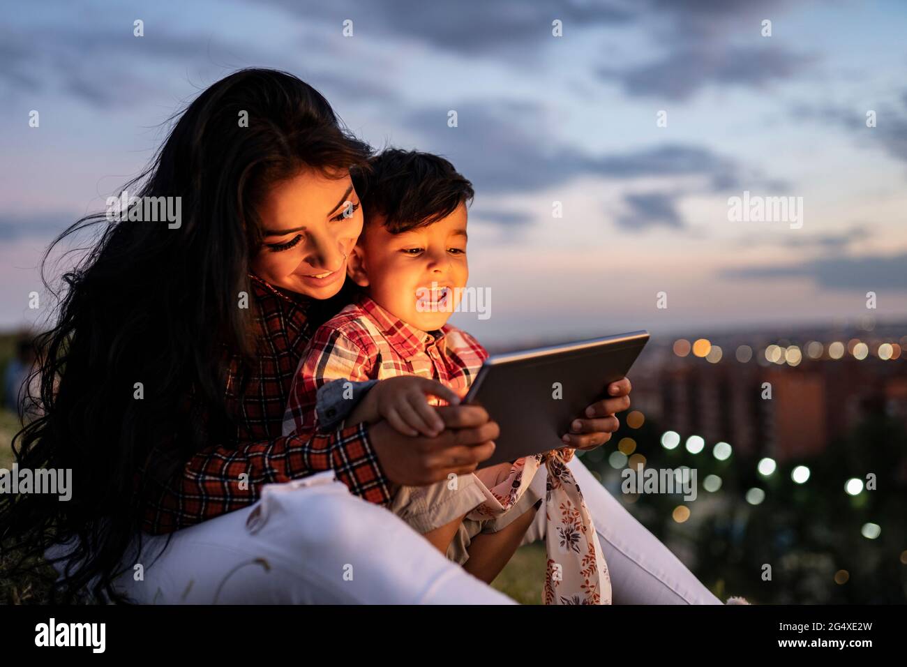Happy son using digital tablet while sitting with mother during sunset Stock Photo