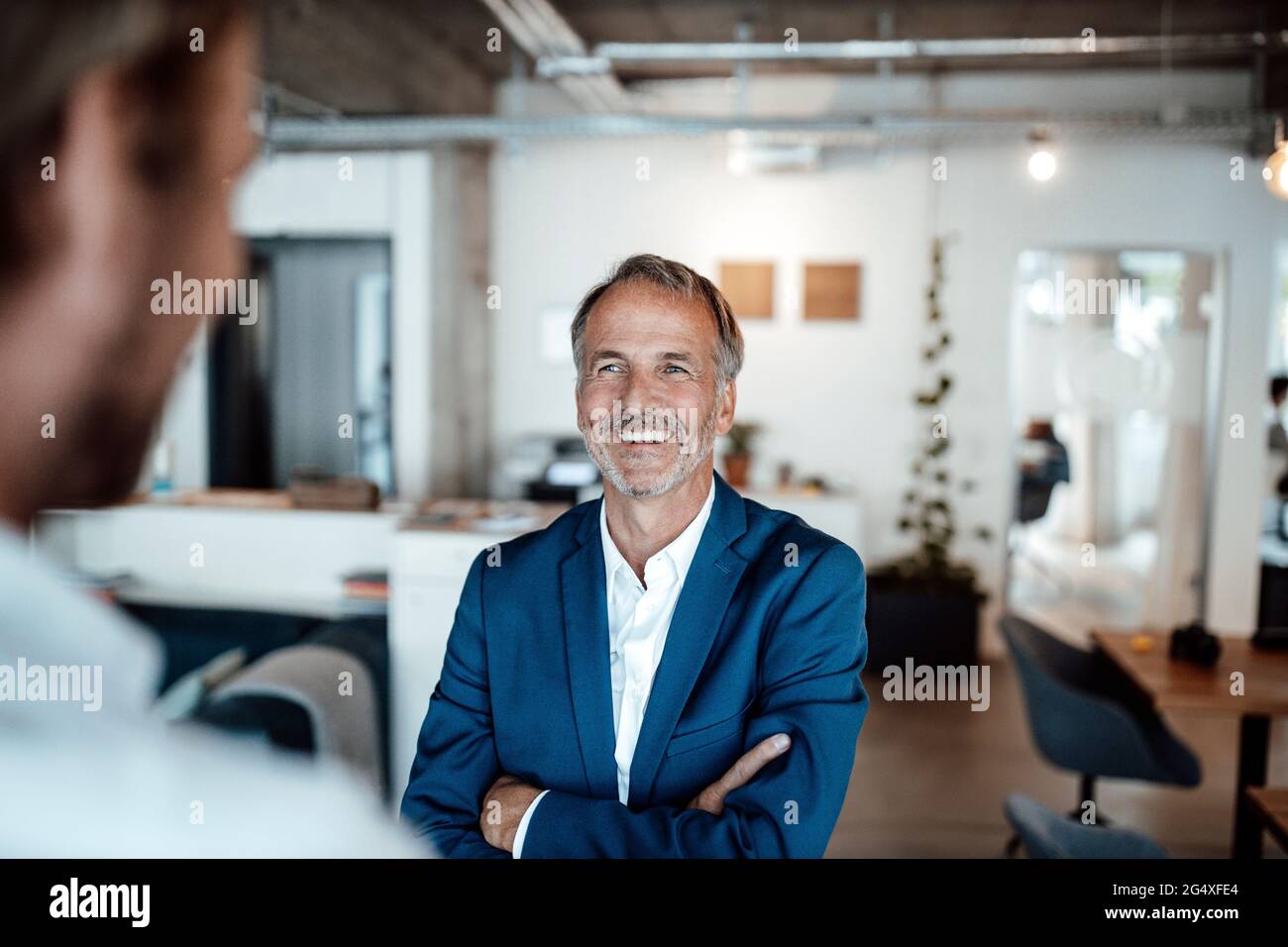 Smiling businessman with arms crossed looking at colleague in office ...