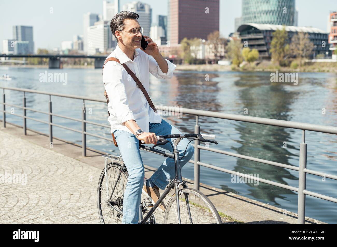 Smiling businessman cycling while talking on mobile phone by river in city Stock Photo