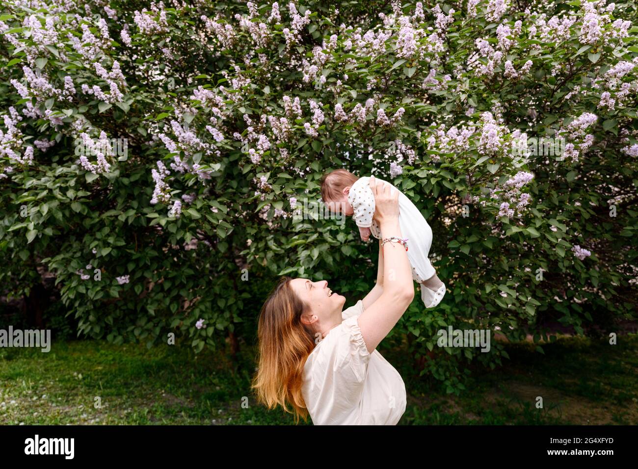 Mother picking up baby girl by flowering plant at park Stock Photo