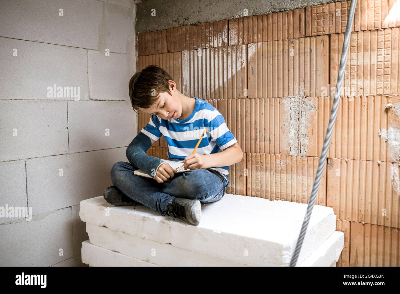 Boy with broken arm writing in book while sitting on block during renovation Stock Photo