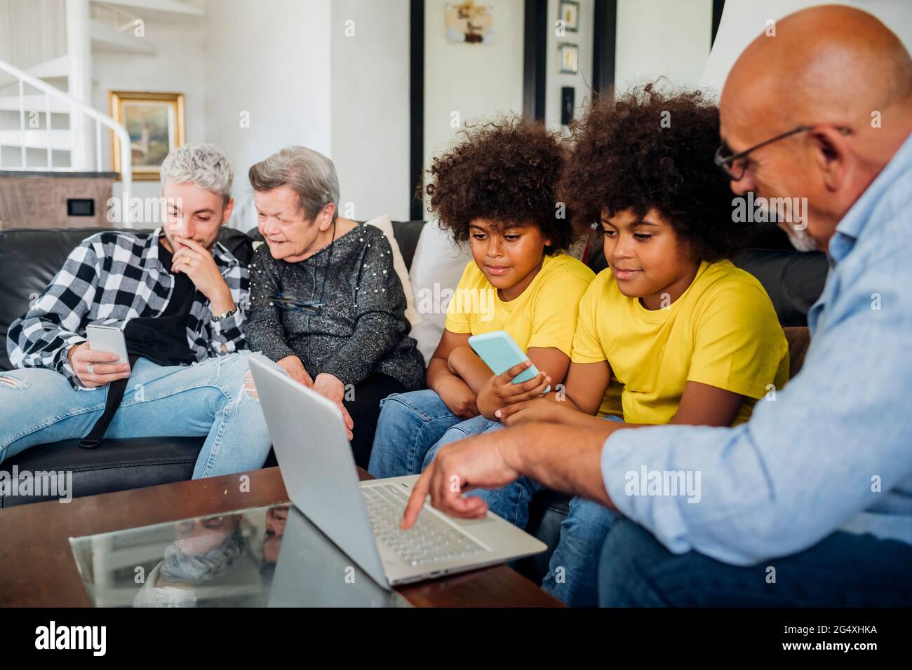 Twin boys with grandfather using laptop while young man using smart phone sitting by great grandmother in living room Stock Photo