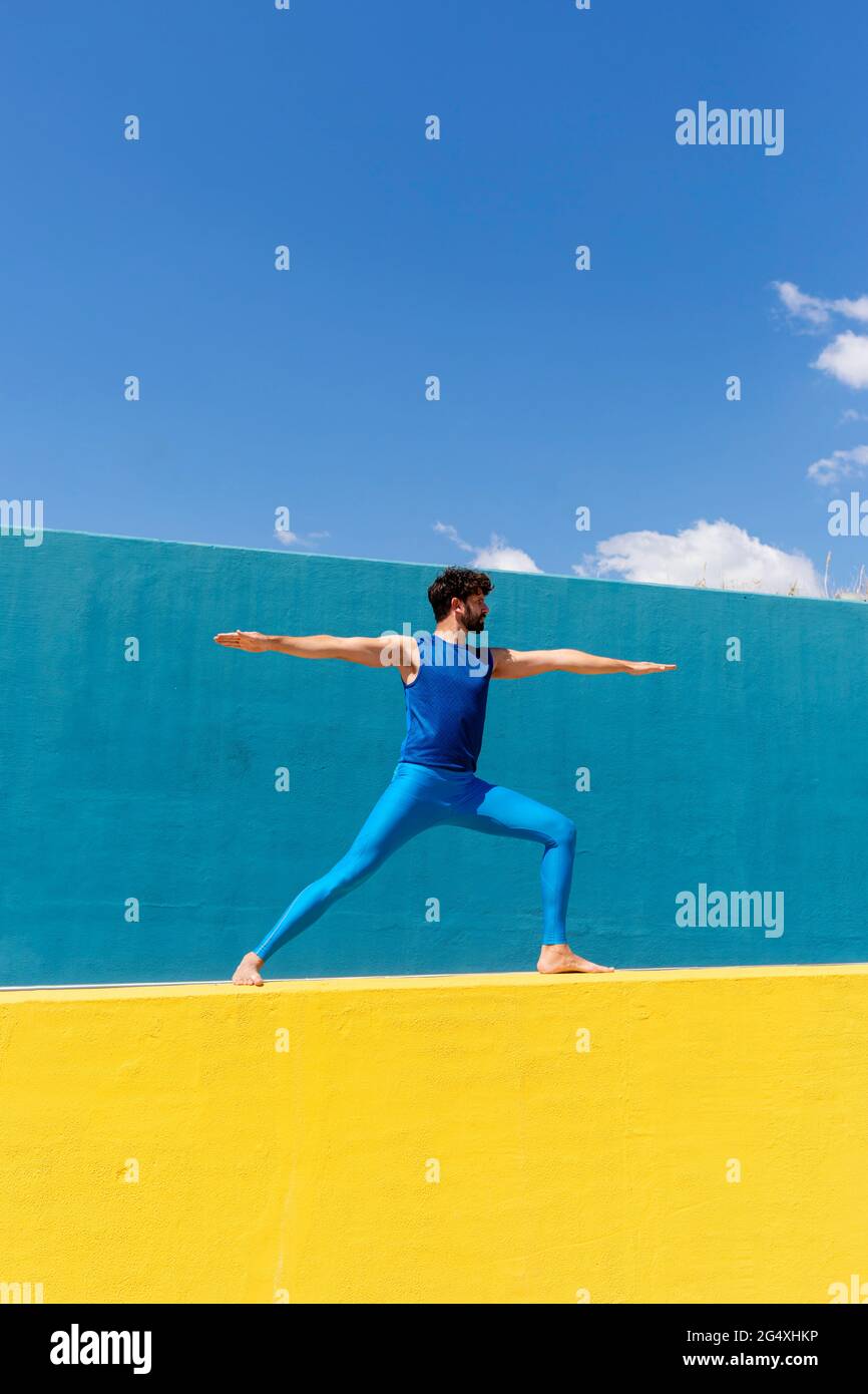Man practicing Virabhadrasana II while doing yoga on retaining wall Stock Photo