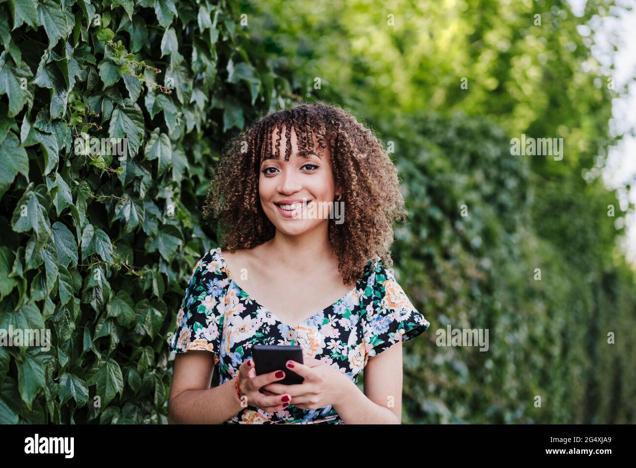 Smiling young woman holding smart phone while standing by ivy plant Stock Photo