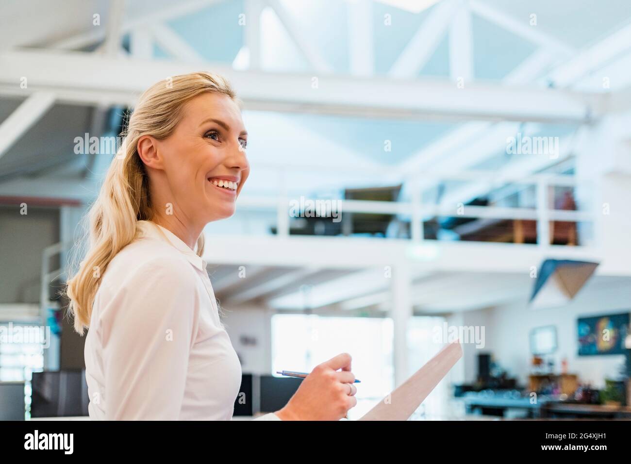 Smiling blond female professional with documents in creative office looking away Stock Photo