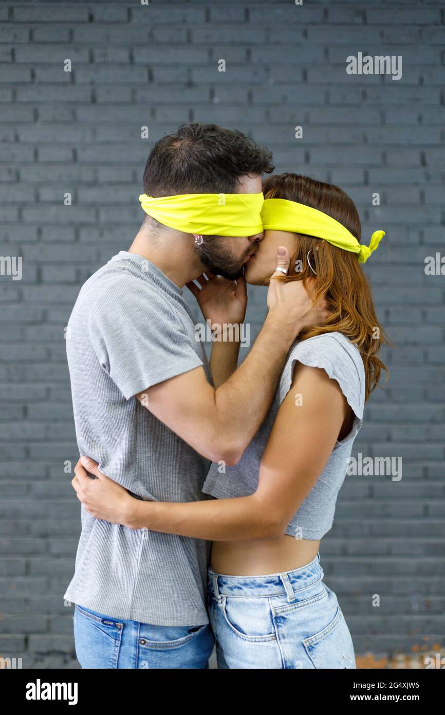 Affectionate couple with eyes covered with cloth kissing each other by gray wall Stock Photo