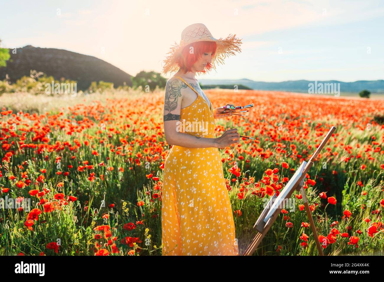Mid adult female artist in yellow sundress painting on poppy field Stock Photo
