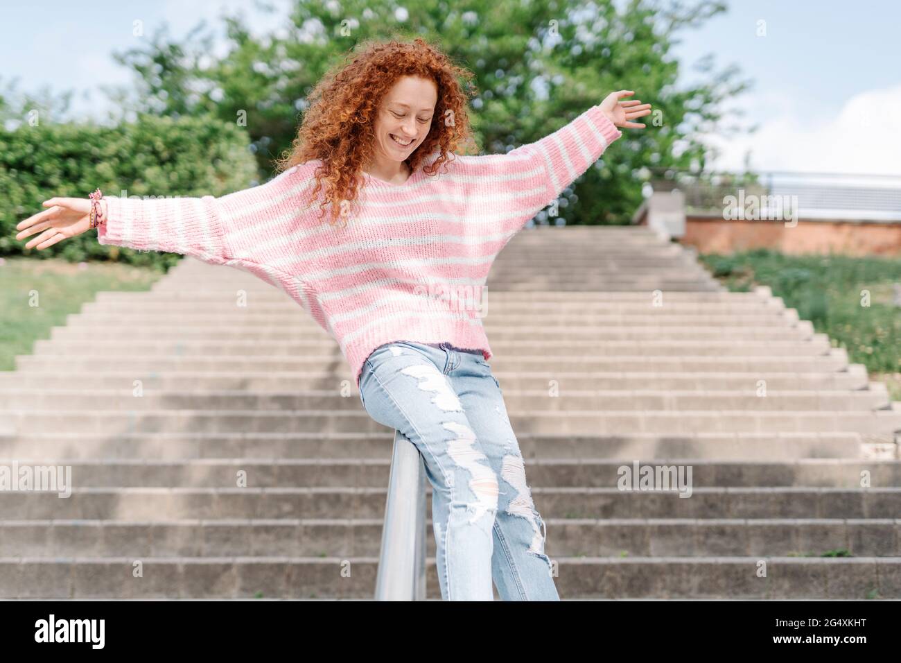 Happy redhead woman with arms outstretched sliding down on railing at park Stock Photo