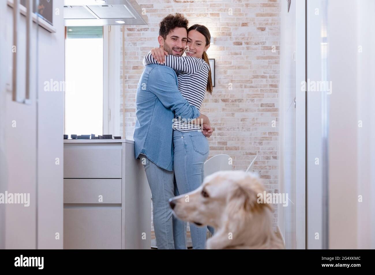 Smiling embracing couple looking at dog in domestic kitchen at home Stock Photo