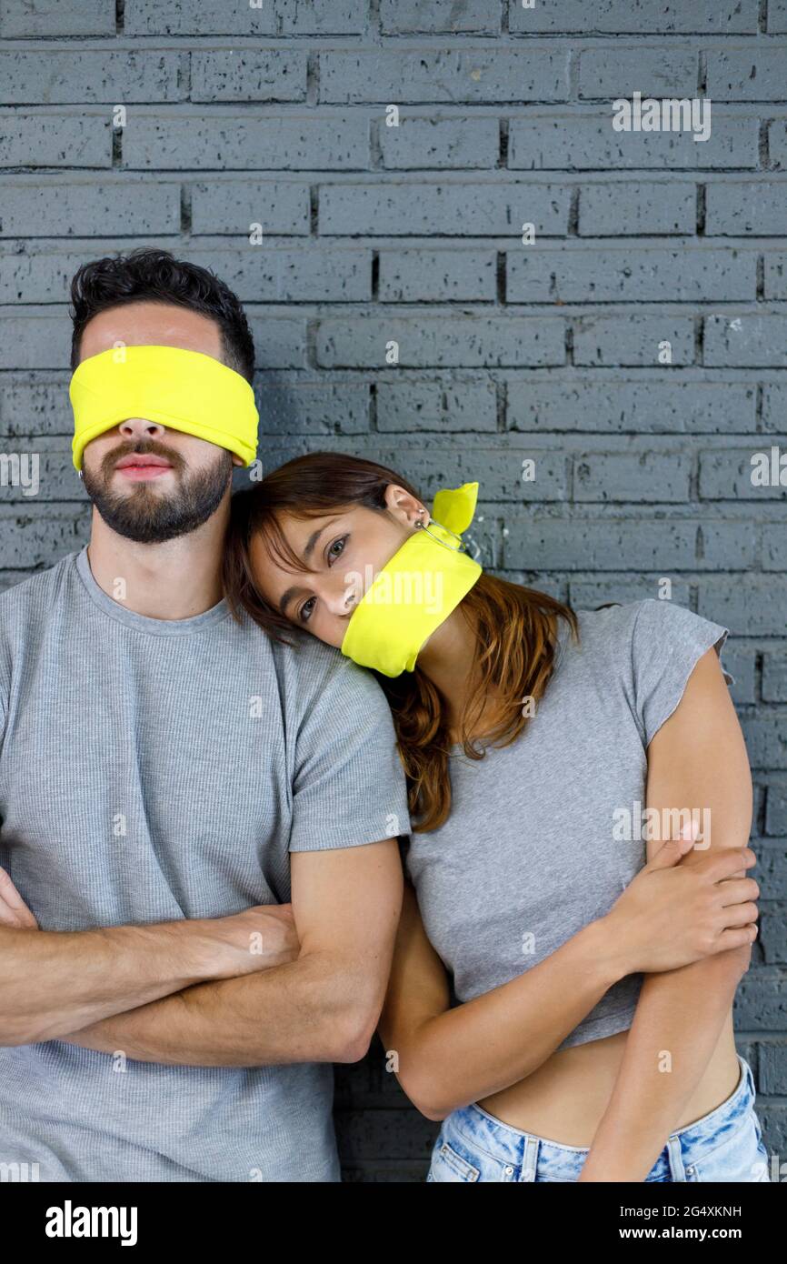 Young couple with mouth and eyes covered with cloth standing in front of brick wall Stock Photo