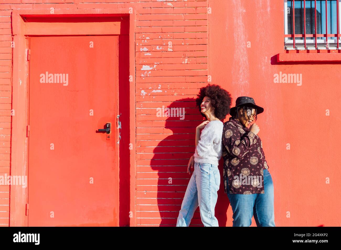 Smiling female friends standing back to back by red building Stock Photo
