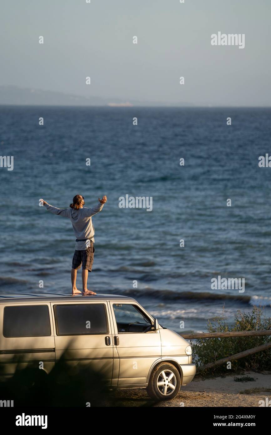 Young man with arms outstretched standing on van roof by sea Stock Photo