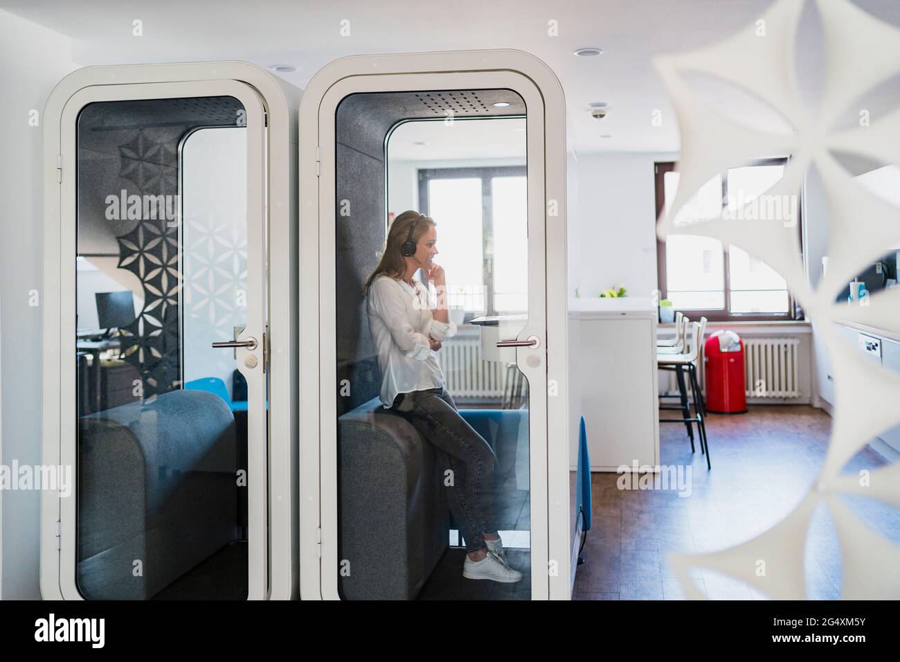 Businesswoman wearing headphones leaning in soundproof cabin at office ...