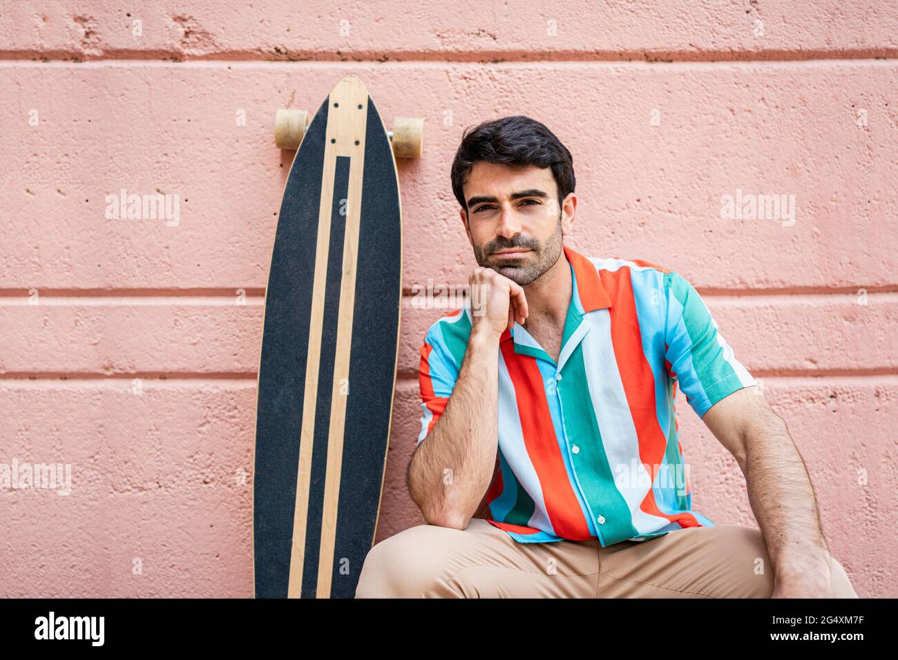 Handsome man with hand on chin sitting by longboard Stock Photo
