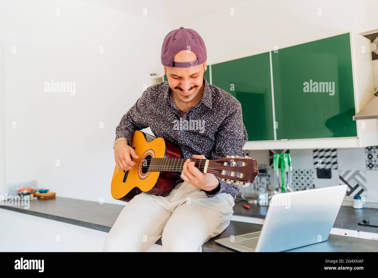 Smiling male composer playing guitar by laptop on kitchen island at home Stock Photo