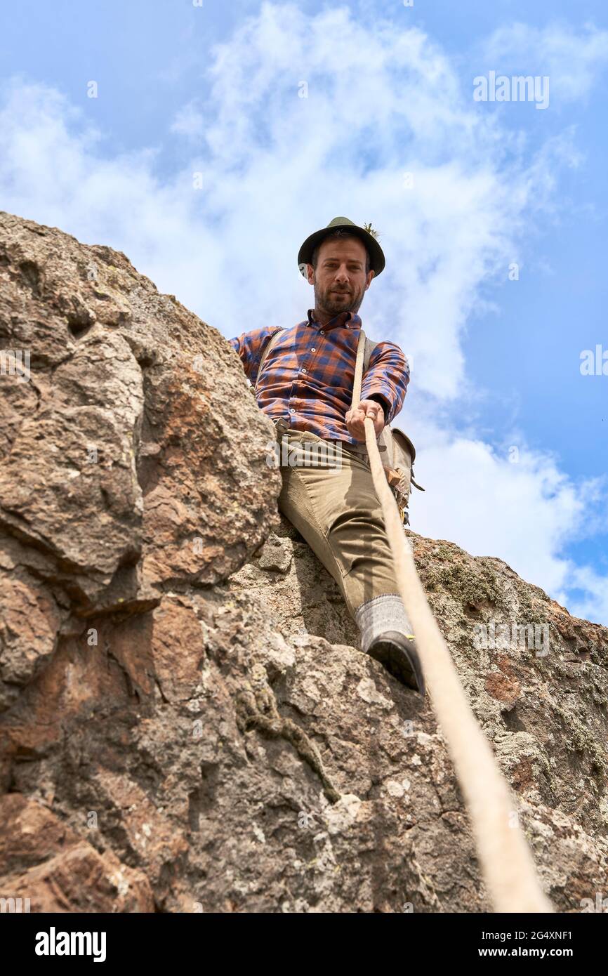 Male mountaineer holding rope while sitting on rock during sunny day Stock Photo