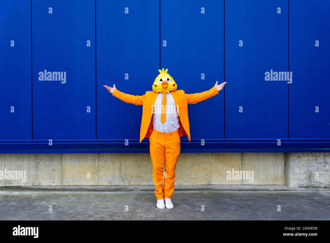 Man wearing vibrant orange suit and bird mask standing in front of blue