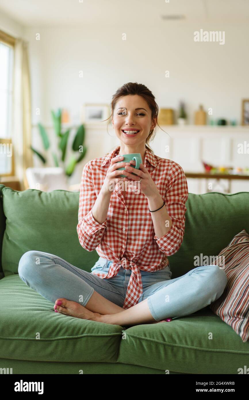 Happy young woman sitting on a couch at home. The woman is drinking tea. Stock Photo