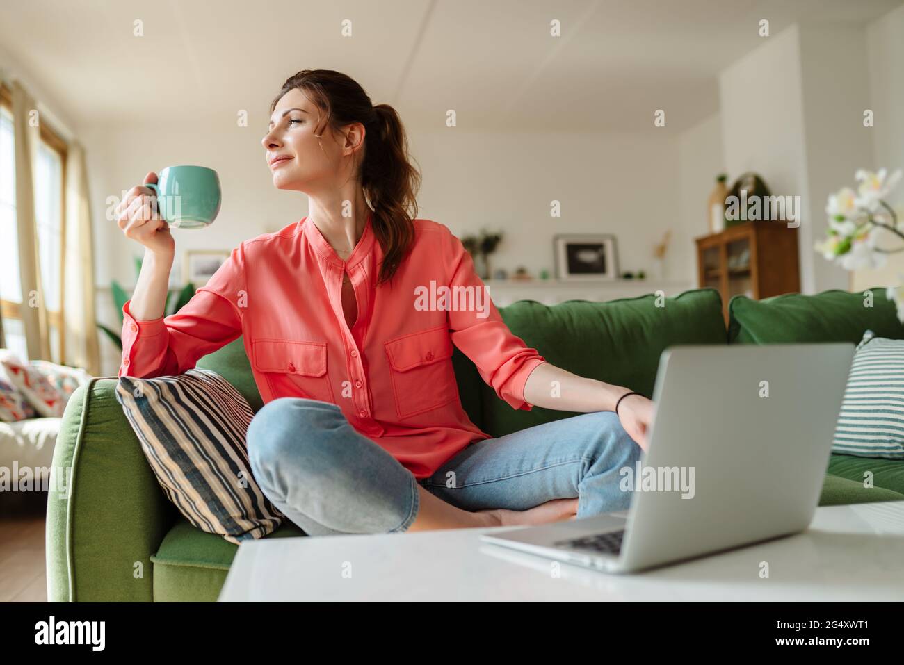 Happy young woman sitting on a couch at home. The woman is drinking tea. Stock Photo