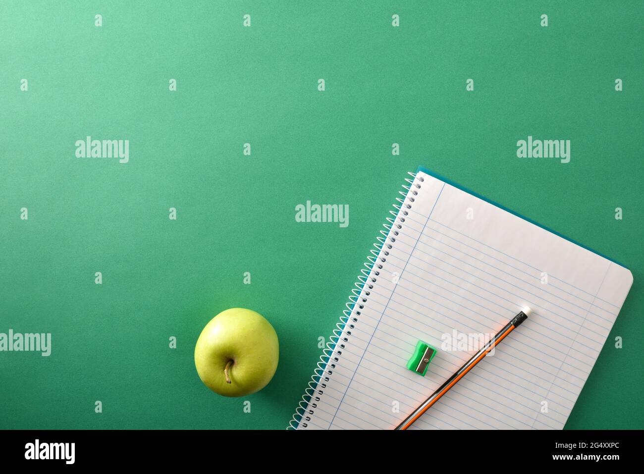 Open spiral notebooks with pencil and sharpener on top and green apple aside on green background with space for text. Top view. Stock Photo