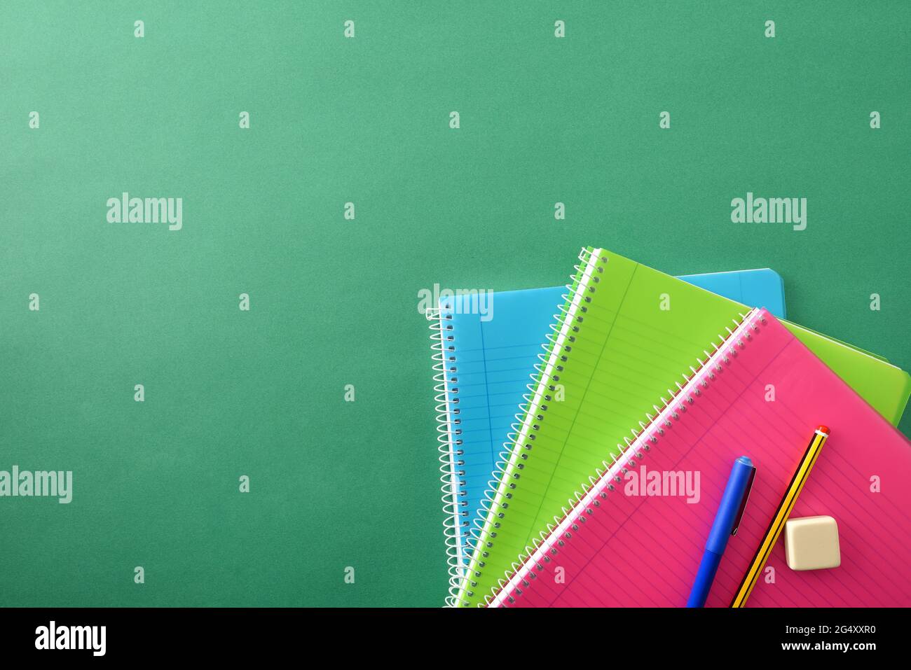 Three colored spiral notebooks with pencil and eraser pen on top on green background with space for text. Top view. Stock Photo