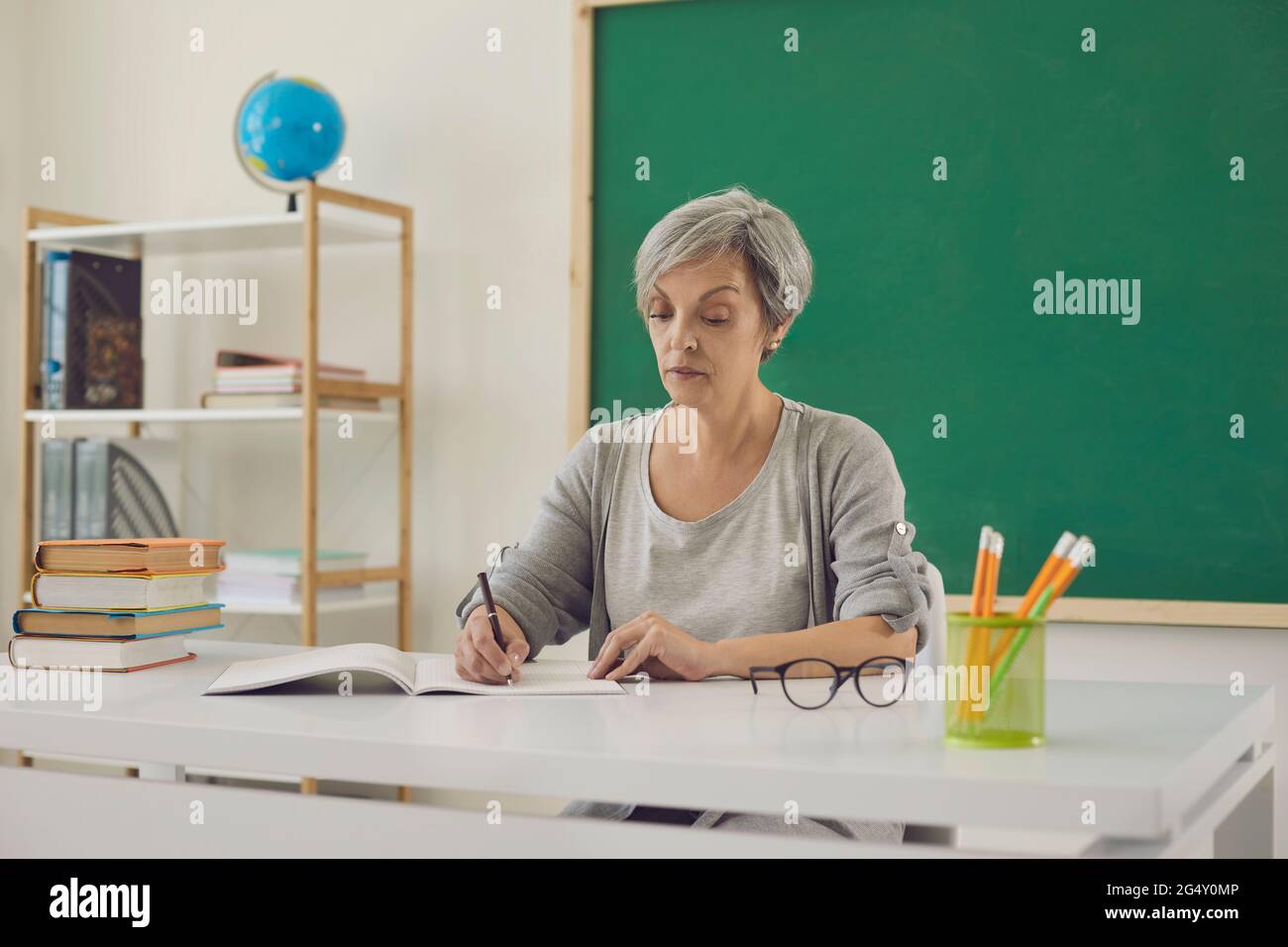 Woman teacher writes while sitting at desk in classroom with green board at school. Education teaching. Stock Photo