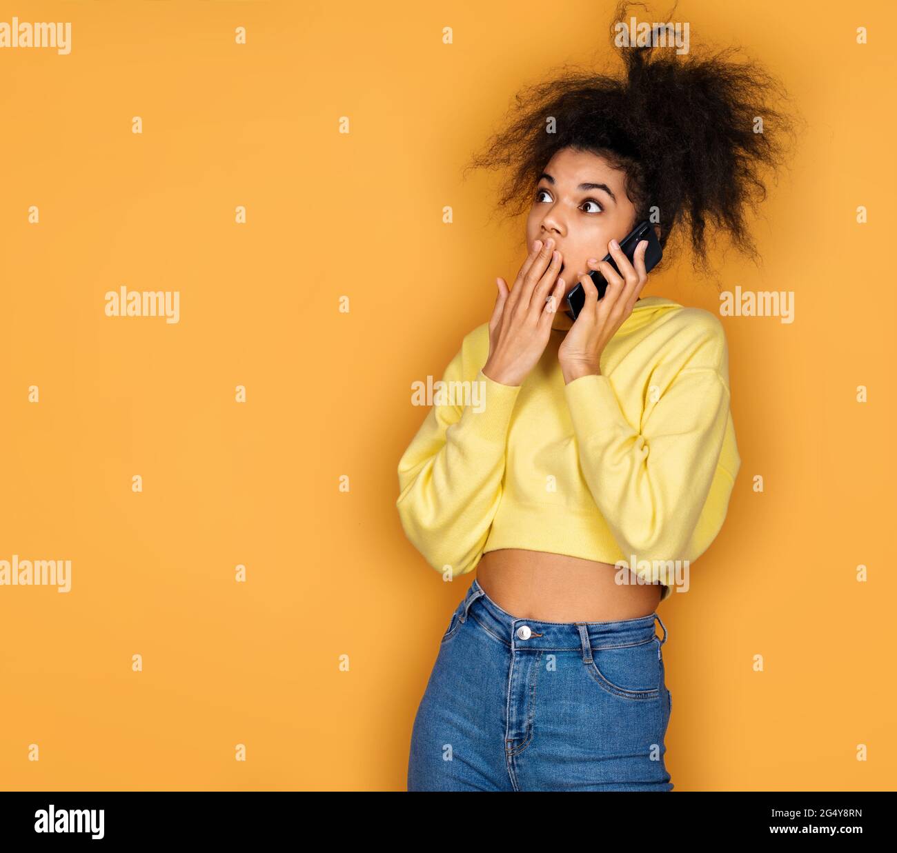 Girl is talking on the phone and looking shocked. Photo of african american girl on yellow background Stock Photo
