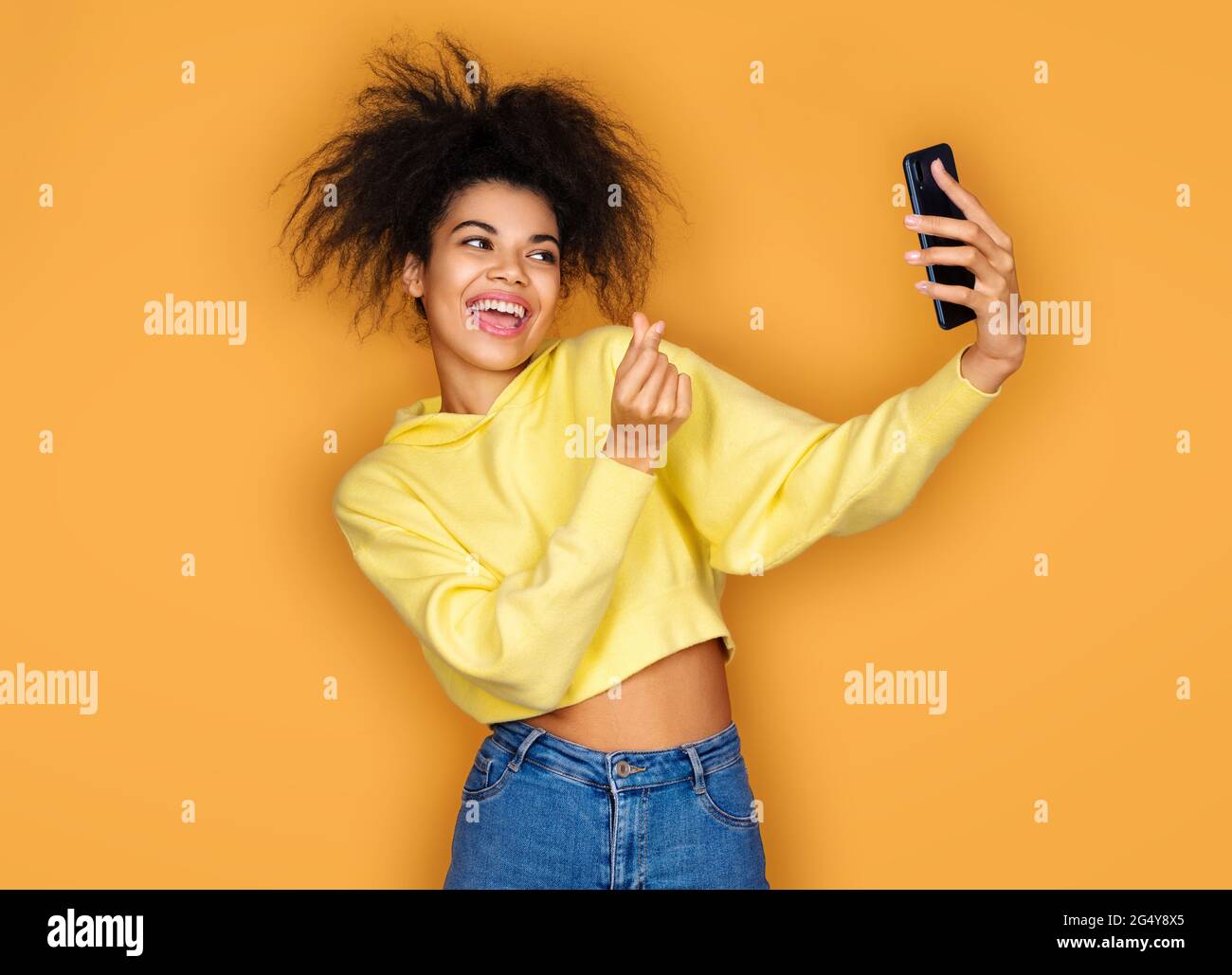 Young girl makes korean love sign and takes selfie photo, looking at phone. Photo of african american girl on yellow background Stock Photo