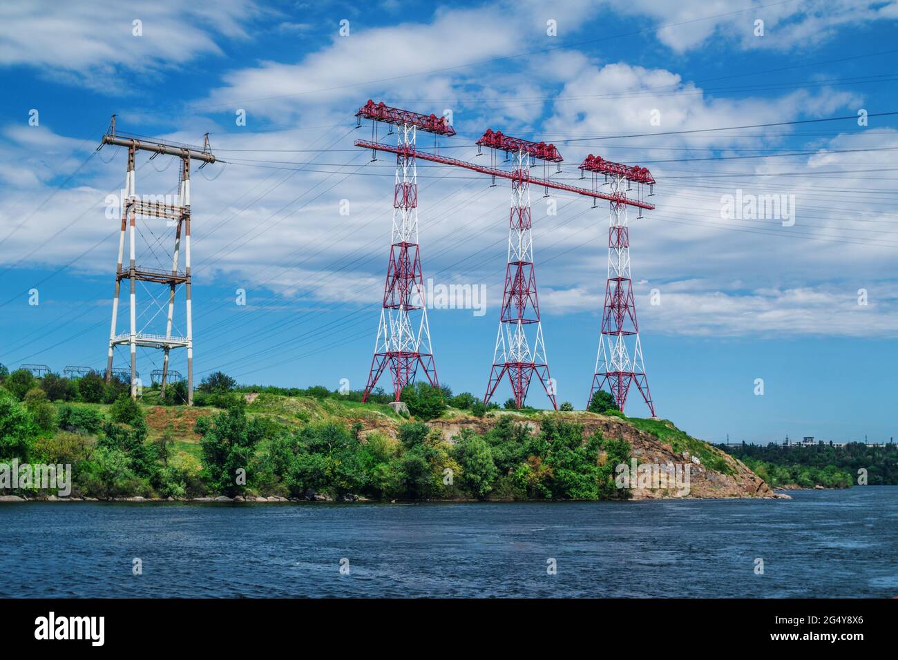 Electricity transmission towers on the river bank. Summer landscape with high voltage post, green trees and blue sky. Stock Photo