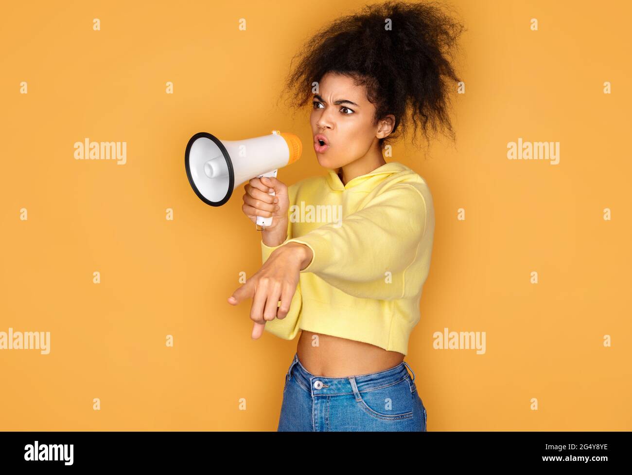 Girl shouts in megaphone and points forefinger to the side. Photo of african american girl on yellow background Stock Photo