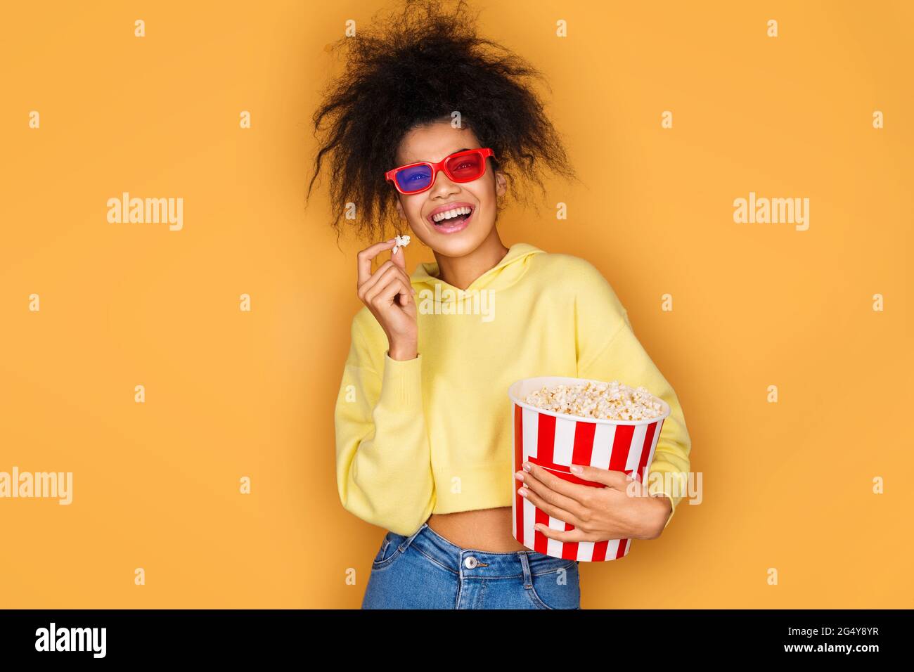 Young girl watching tv with popcorn box. Photo of african american girl on yellow background Stock Photo