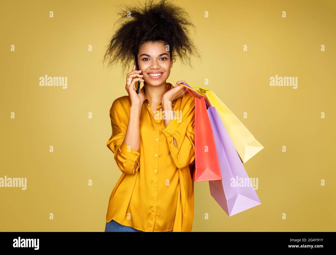 Happy girl holds shopping bags and talks by mobile phone. Photo of african american girl on yellow background Stock Photo