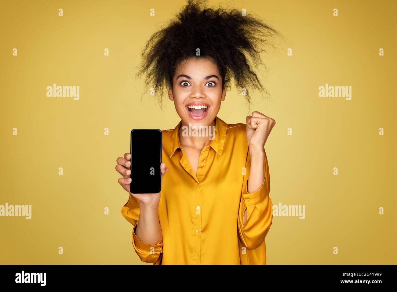 Excited girl shows smartphone and clenches fist, feels overjoyed after winning or good news. Photo of african american girl on yellow background Stock Photo
