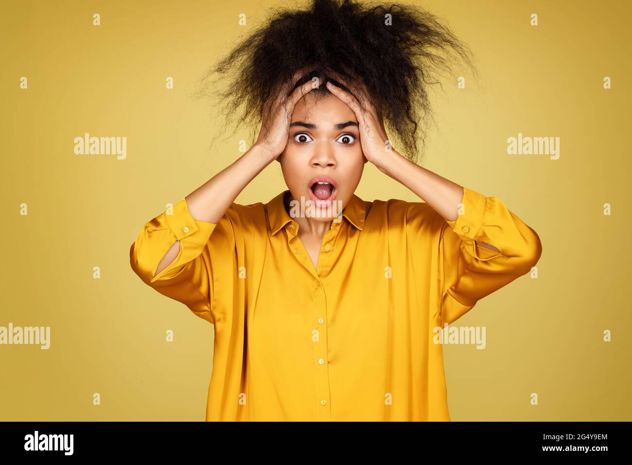 Shocked girl holds hands on head, stares in terror. Photo of african american girl on yellow background Stock Photo
