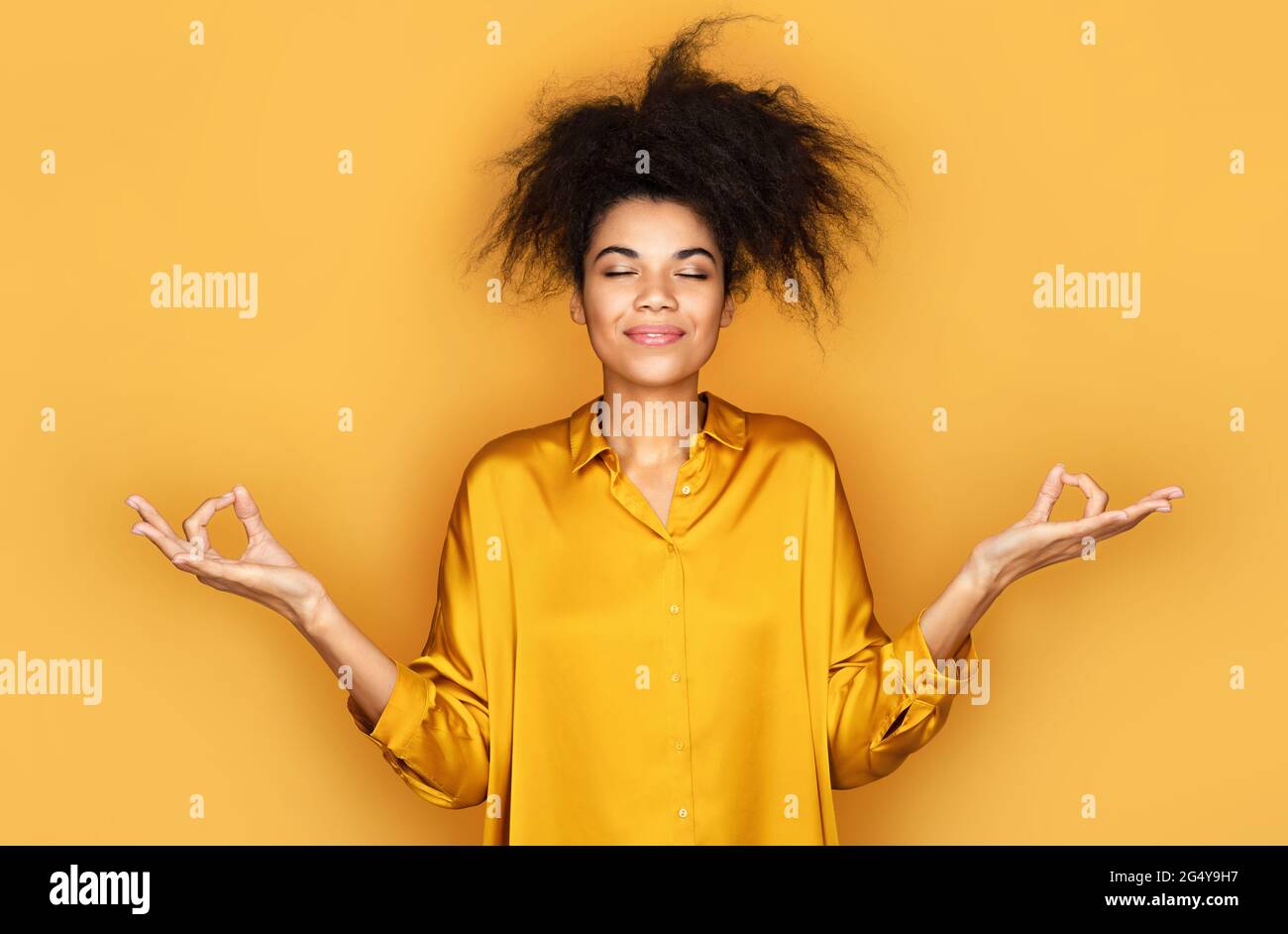 Relaxed girl with closed eyes, holds hands in zen gesture. Photo of african american girl on yellow background Stock Photo