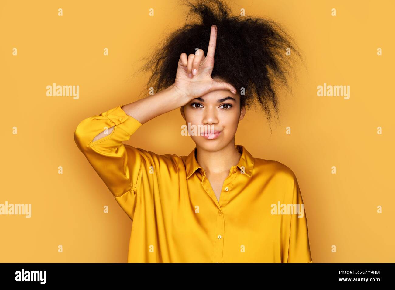 Girl making fun of people with fingers on forehead doing loser gesture mocking and insulting. Photo of african american girl on yellow background Stock Photo