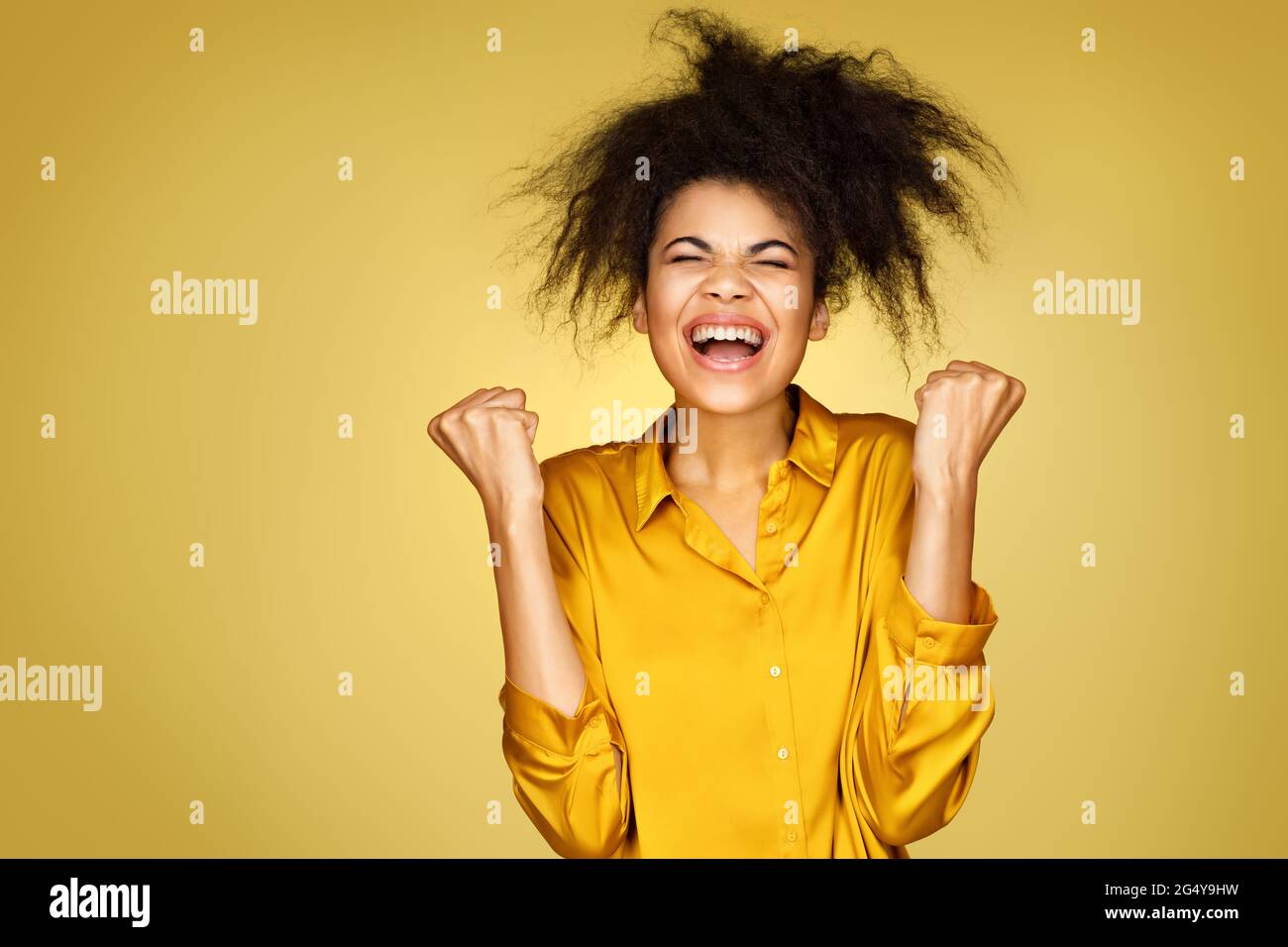 Joyful girl holds fists clenched, celebrates success. Photo of african american girl on yellow background Stock Photo