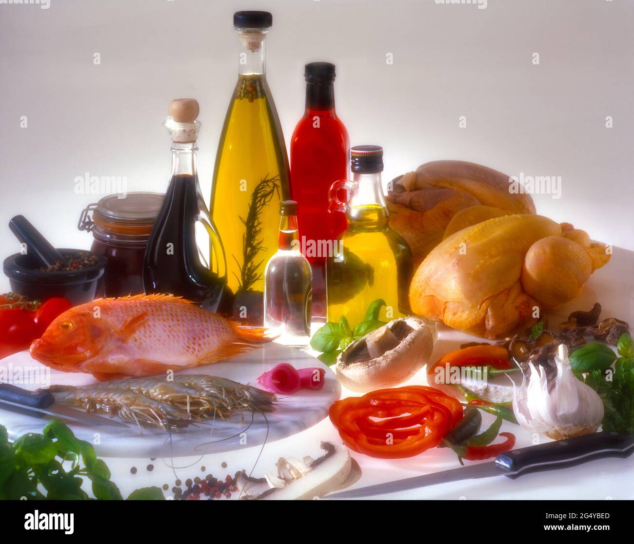Five glass bottles containing coloured liquids raw iingredients poultry, fish prawns garlic mushrooms red peppers pestle & mortar on white copy space Stock Photo