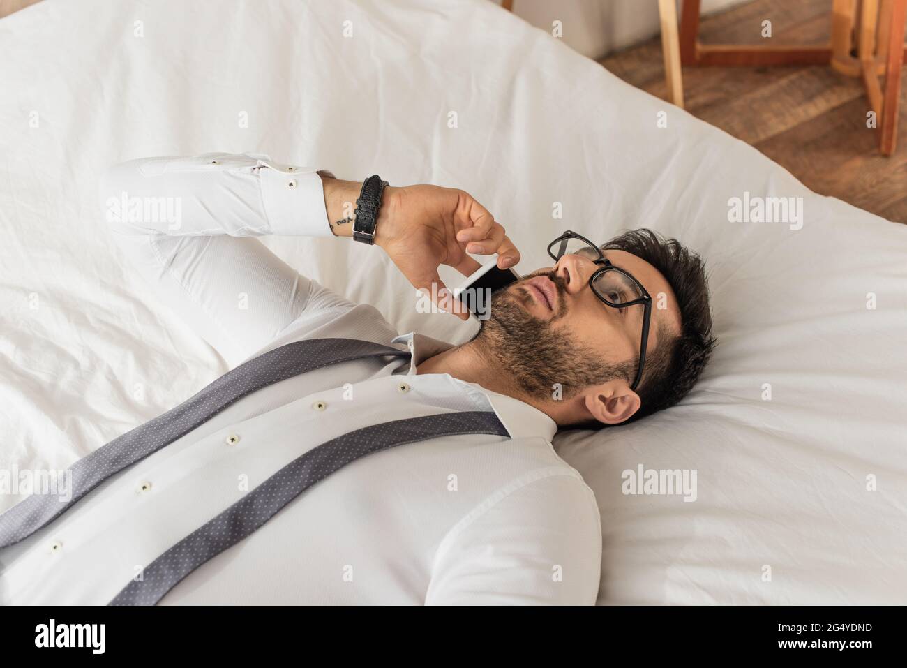 Businessman in eyeglasses and shirt talking on smartphone while lying on bed Stock Photo