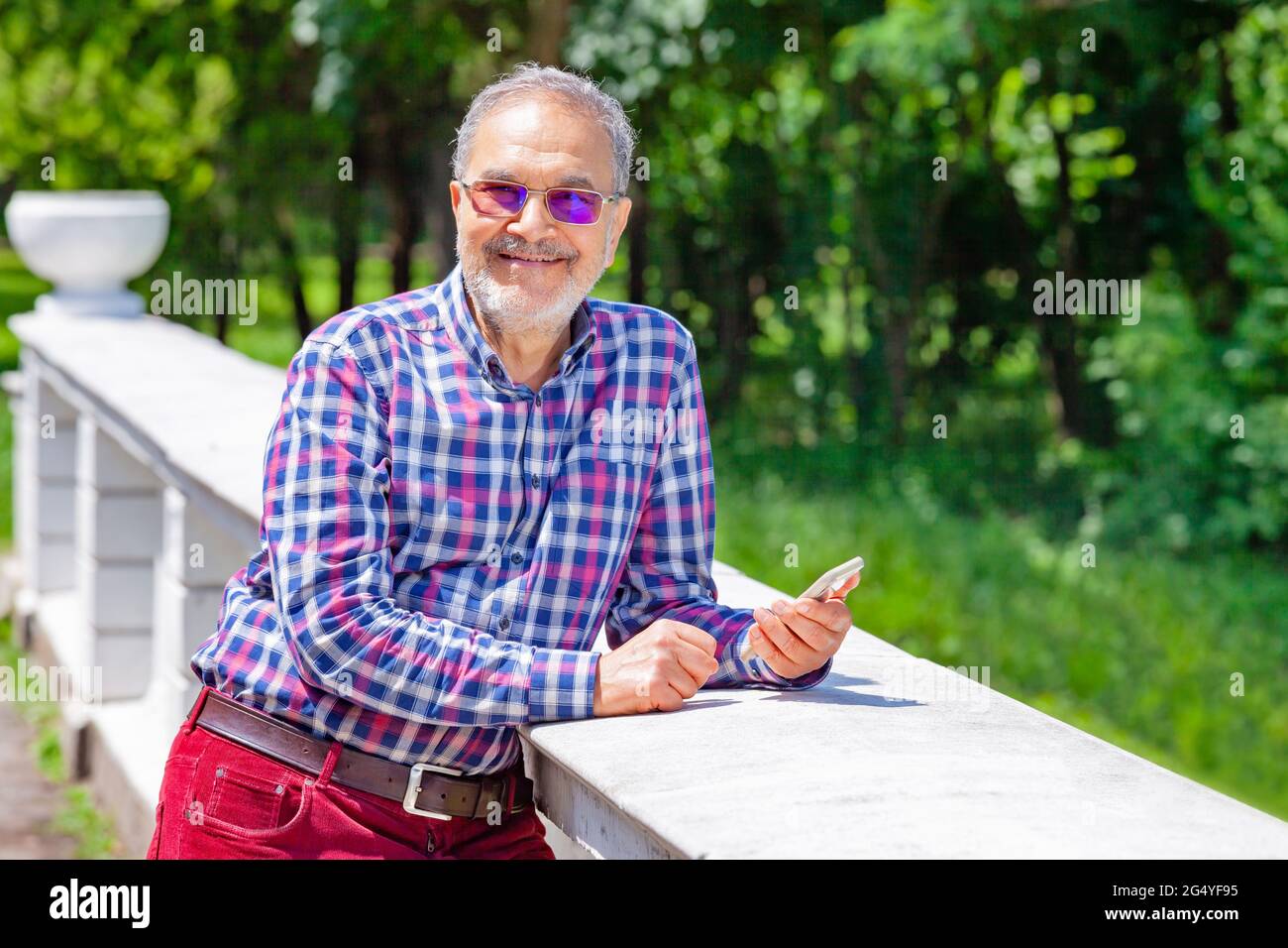 Happy pensioner in his seventies with eye glasses and phone in his hand smiling relaxed in the park during spring or summer time, looking at camera. Stock Photo