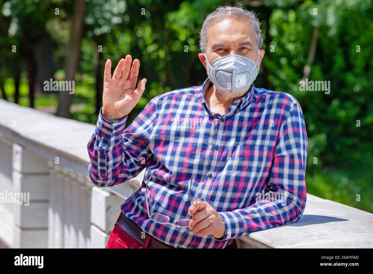 Elderly men with casual clothing in the park during spring or summer time, wearing face protective mask is waving at camera. Stock Photo