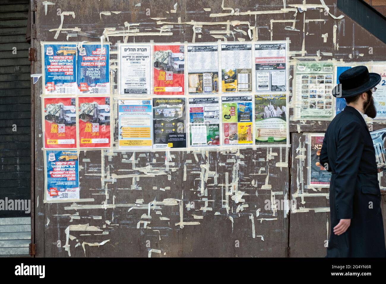 A Hasidic Jew passes a wall of adverts & notices that are primarily in