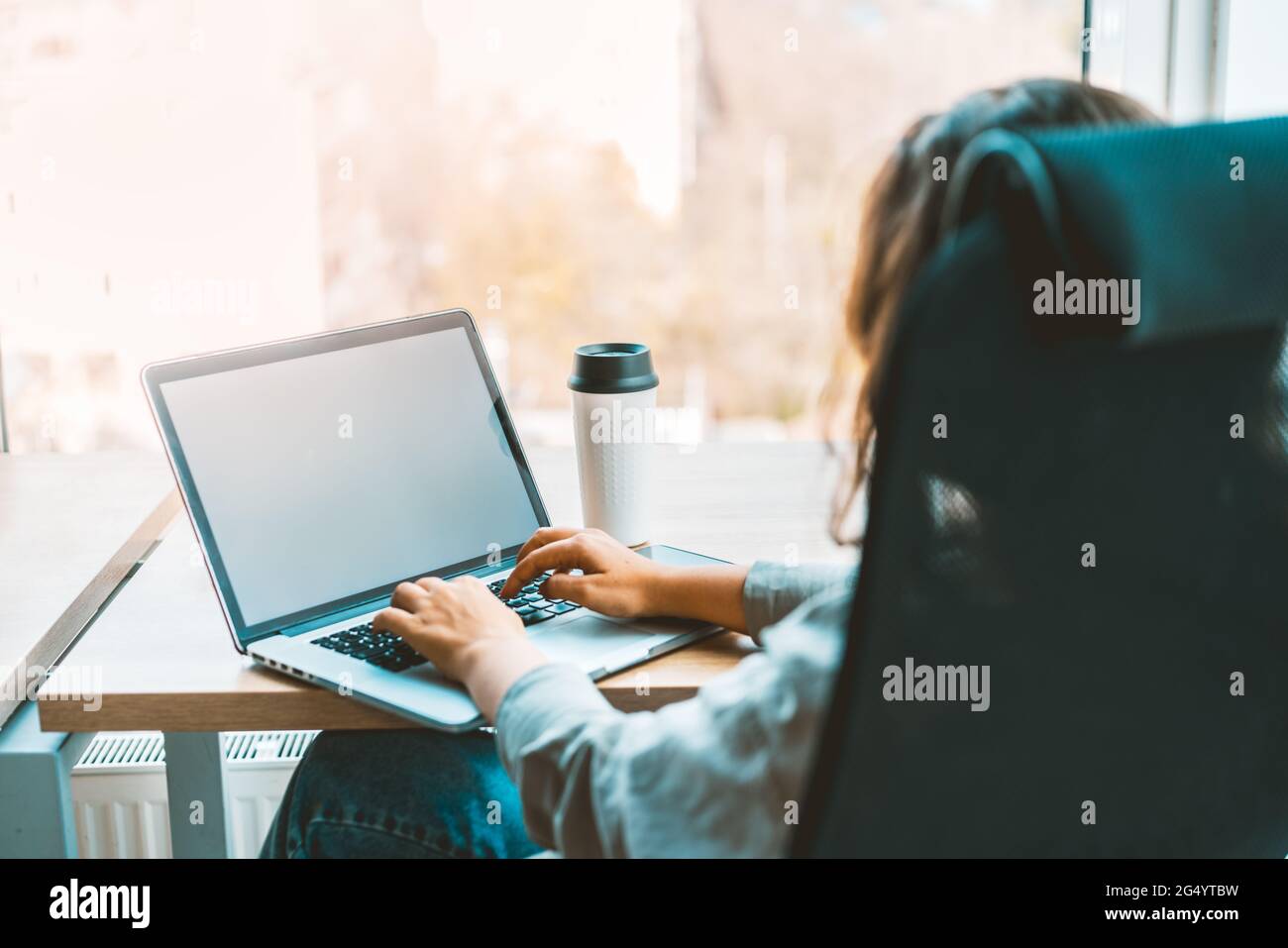 Woman working with a laptop at the modern bright office with a eco-friendly coffee cup, sitting at the desk. Blank white screen for text copy Stock Photo
