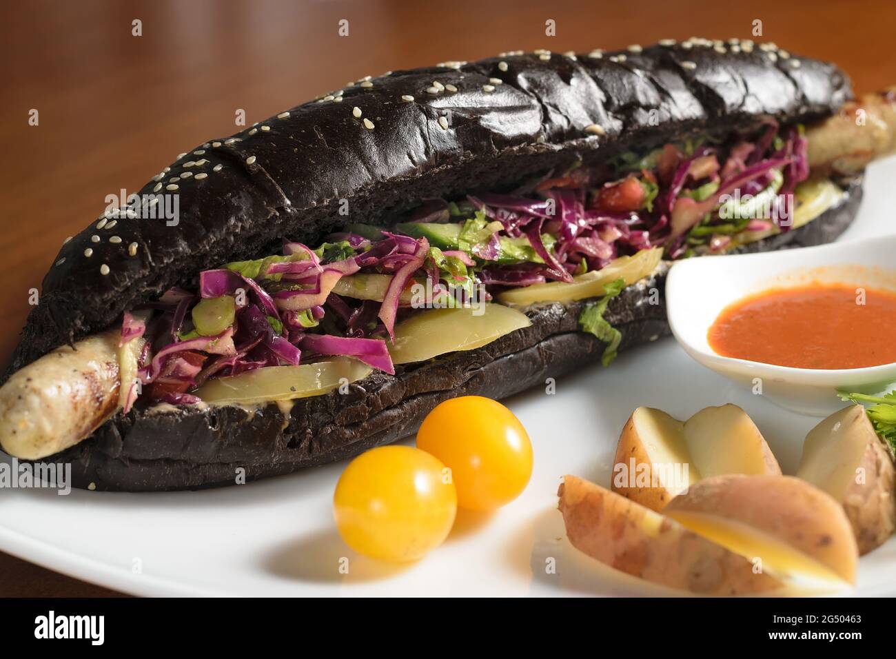 Long black hot dog with fried chicken sausage with salad, on a plate with sauce, tomatoes and potatoes, close-up Stock Photo