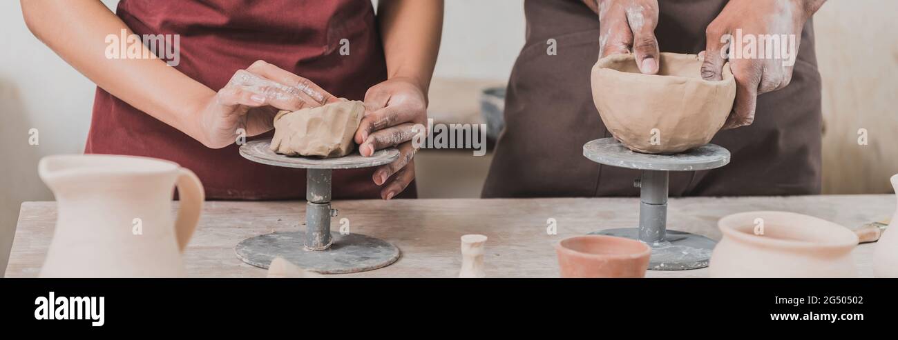partial view of young african american couple in aprons making clay pots on table with equipment in pottery, banner Stock Photo