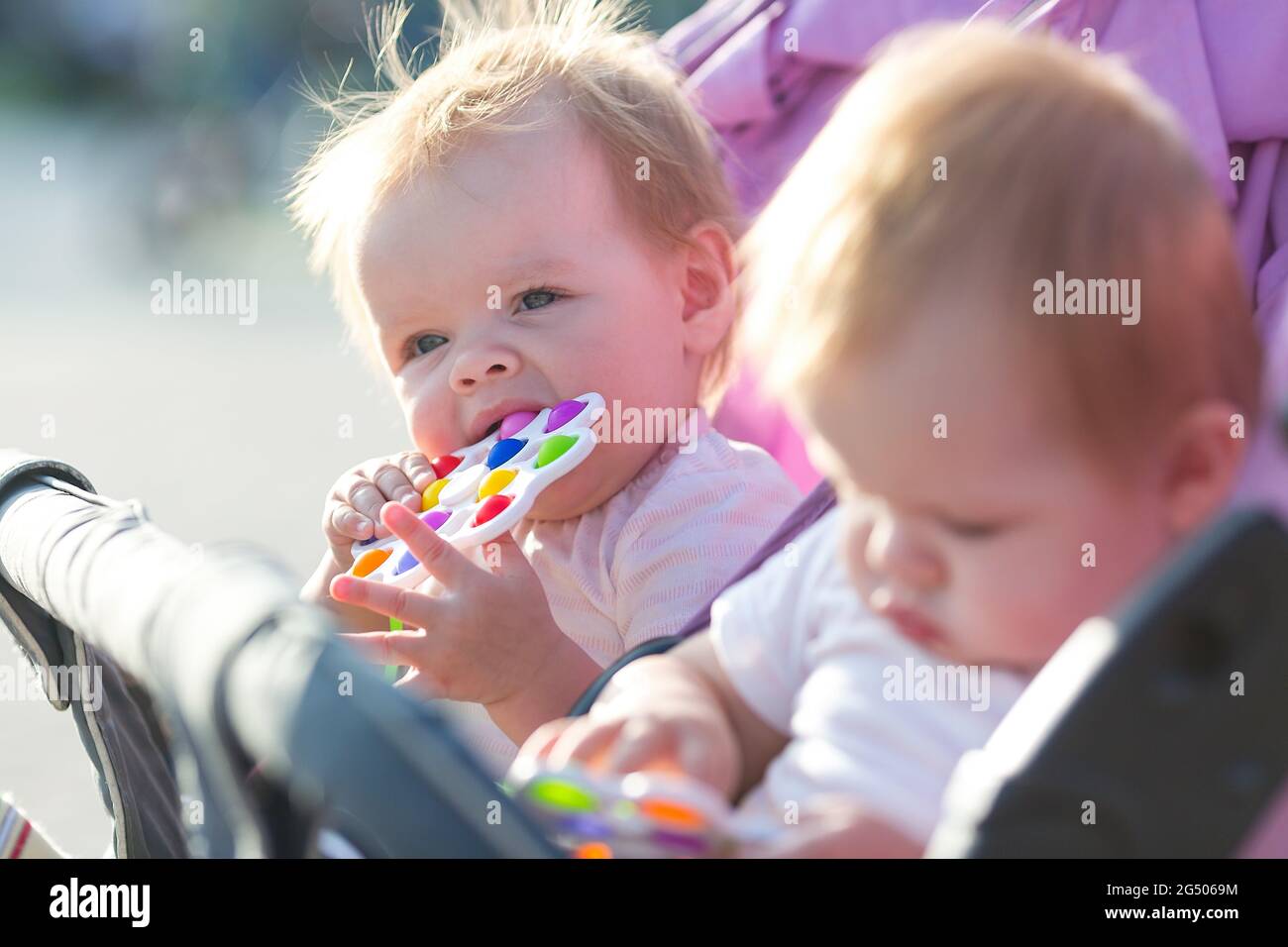 Twin children sit in a baby carriage with toys in their hands. Stock Photo